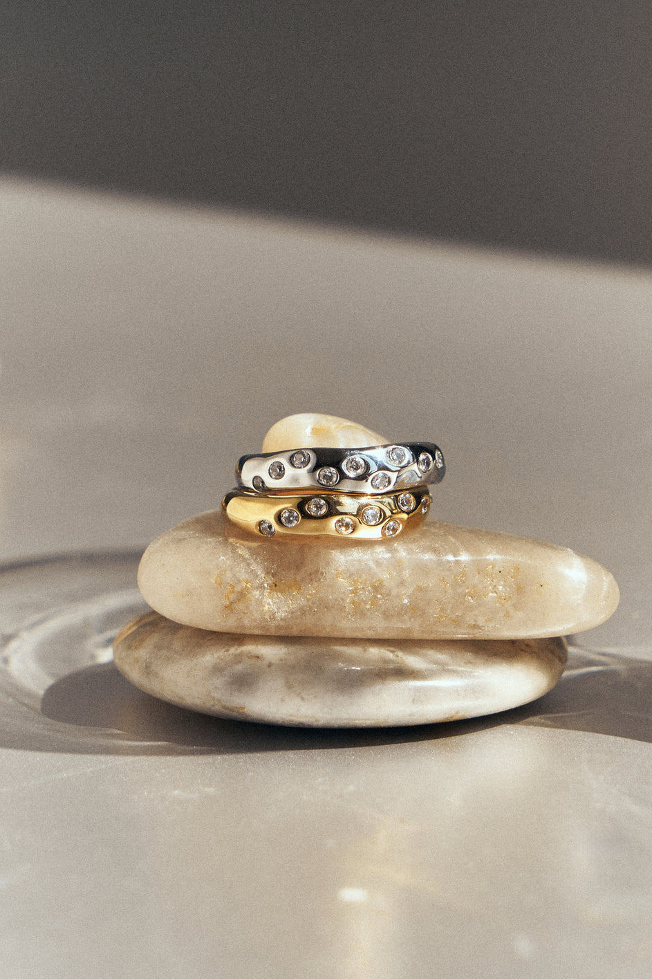 Gold-tone and silver-tone rings with clear stones stacked on smooth pebbles.