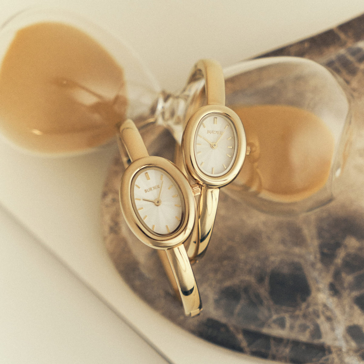 Two gold-tone BURKER watches with oval dials displayed beside coffee cups.