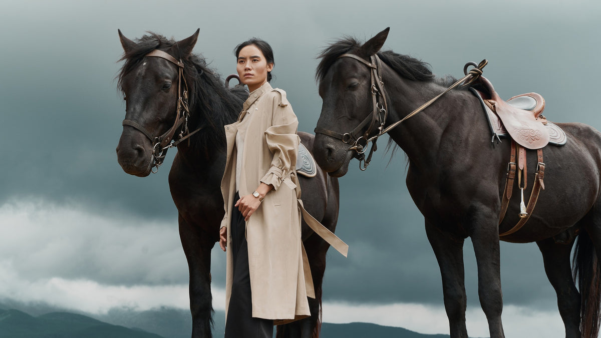 Woman in a beige trench coat wearing a BURKER watch between two black horses.